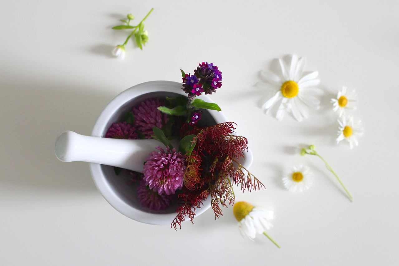 A white mortar and pestle filled with colorful flowers, including purple and red blooms, with scattered white daisies around, symbolizing natural and essential ingredients used in skin care.