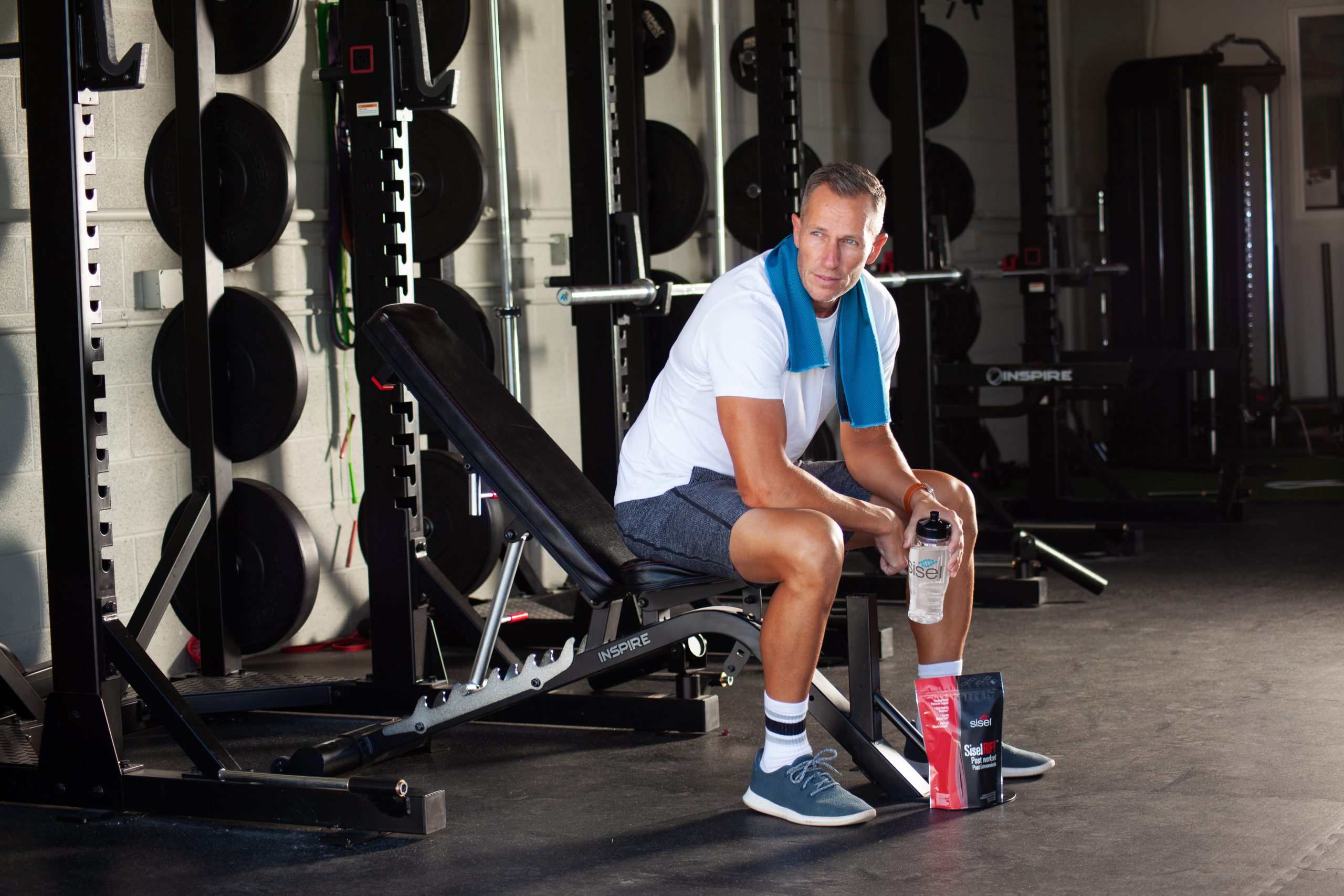 Man sitting on a gym bench with a towel around his neck, holding a shaker bottle and surrounded by clean workout supplements, emphasizing a healthy fitness routine.